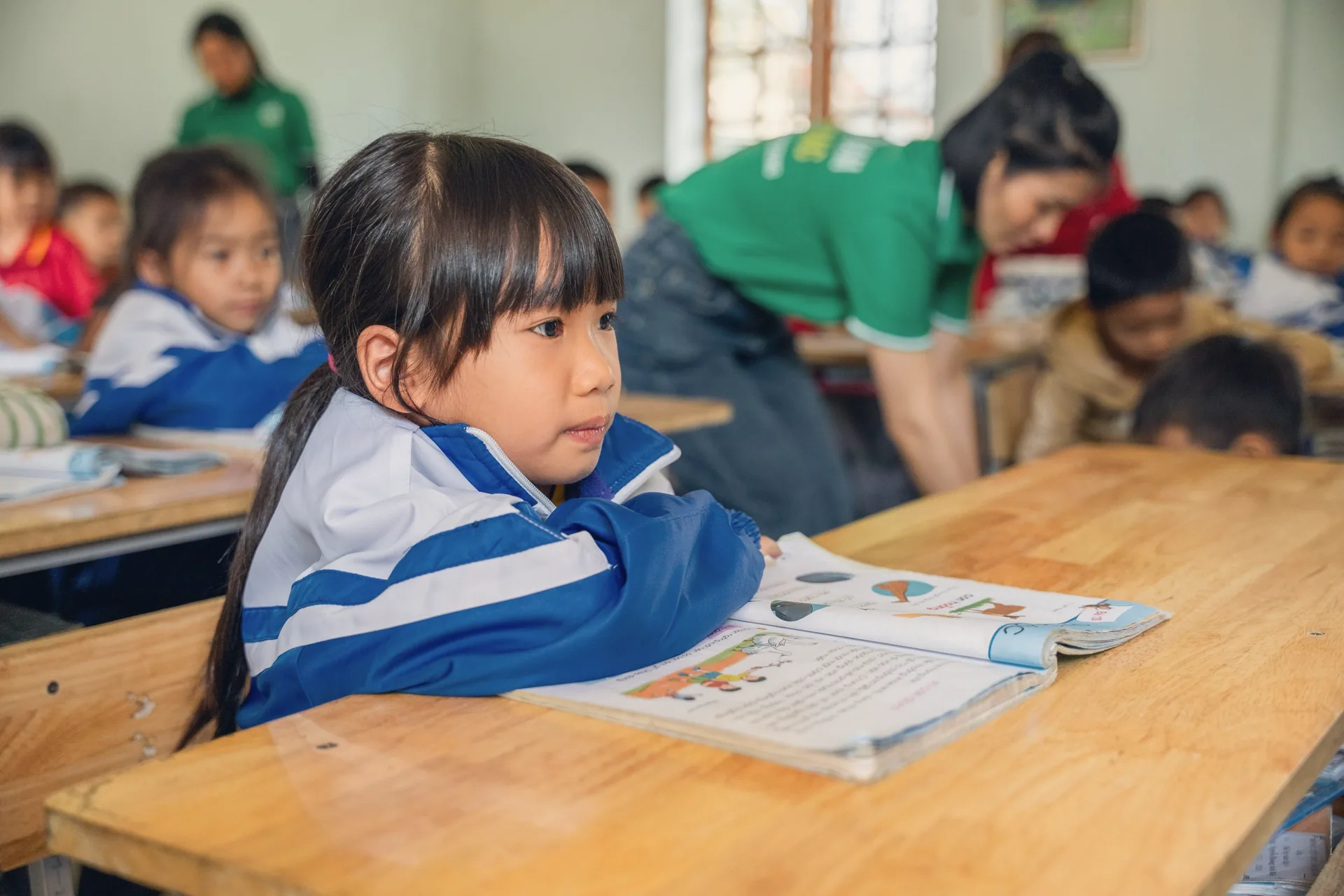 A girl listening at her desk while the teacher helps classmates behind her
